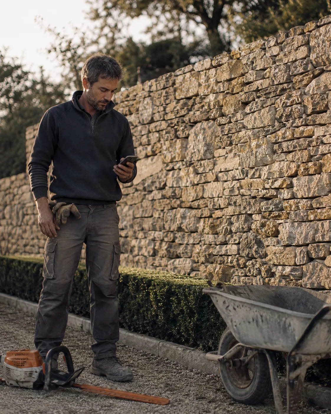 Paysagiste consultant son téléphone devant un mur de pierre, en fin de chantier